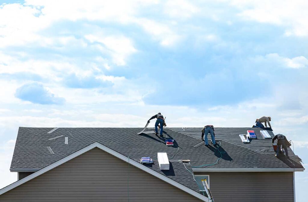 Homeowner compares a new shingle sample with weathered shingles beside a residential roof.